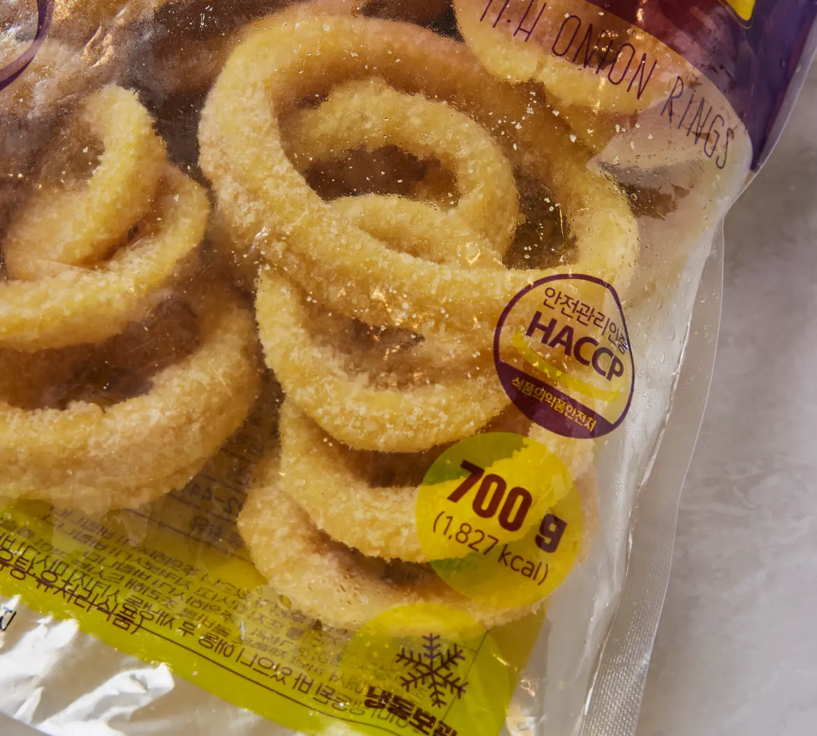 M.H 어니언링 700g 패키지 전면의 투명창으로 보이는 동그란 링 제형과 HACCP 인증 마크 및 중량 칼로리 표시 확대 이미지